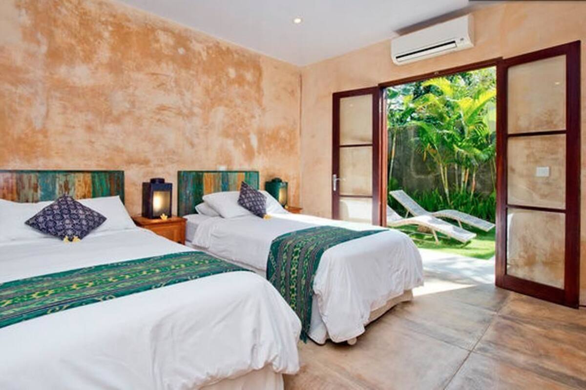 A room with two beds, a large sliding glass door leading to a garden, and a ceiling air conditioning unit.