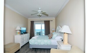 A hotel room with a bed, a desk with a computer, a TV, a ceiling fan, and a view of the ocean through a large window.