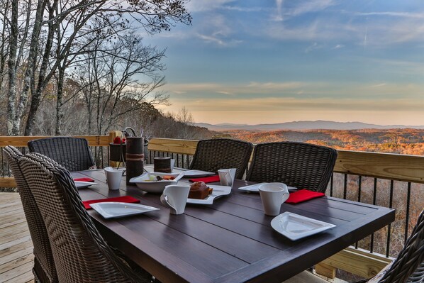 Una mesa de comedor colocada en una terraza de madera con sillas de mimbre, con vistas a un paisaje montañoso.