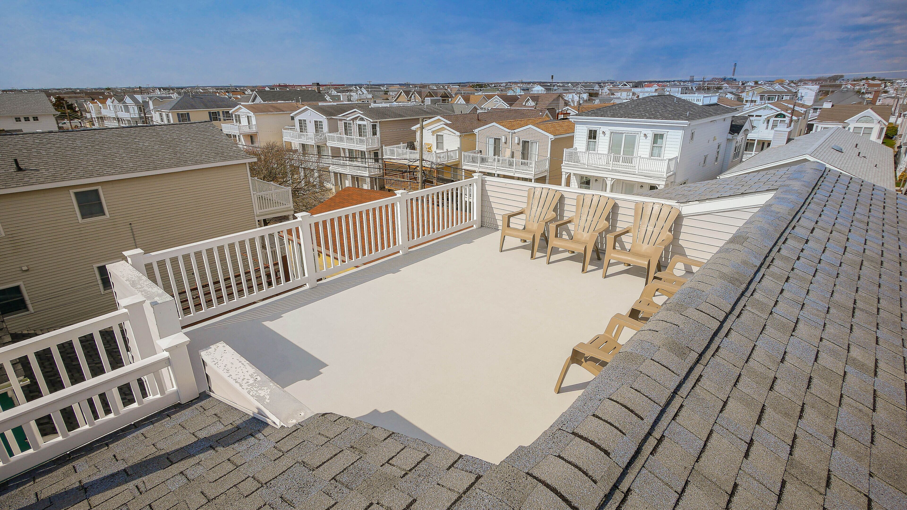 A rooftop with a white railing, beige floor, and several Adirondack chairs.