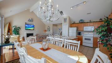 A spacious living area with a wooden dining table, white chairs, a chandelier, and a television mounted on the wall.