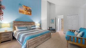 Un dormitorio con cama, mesitas de noche, una silla, una puerta y un cuadro en la pared.