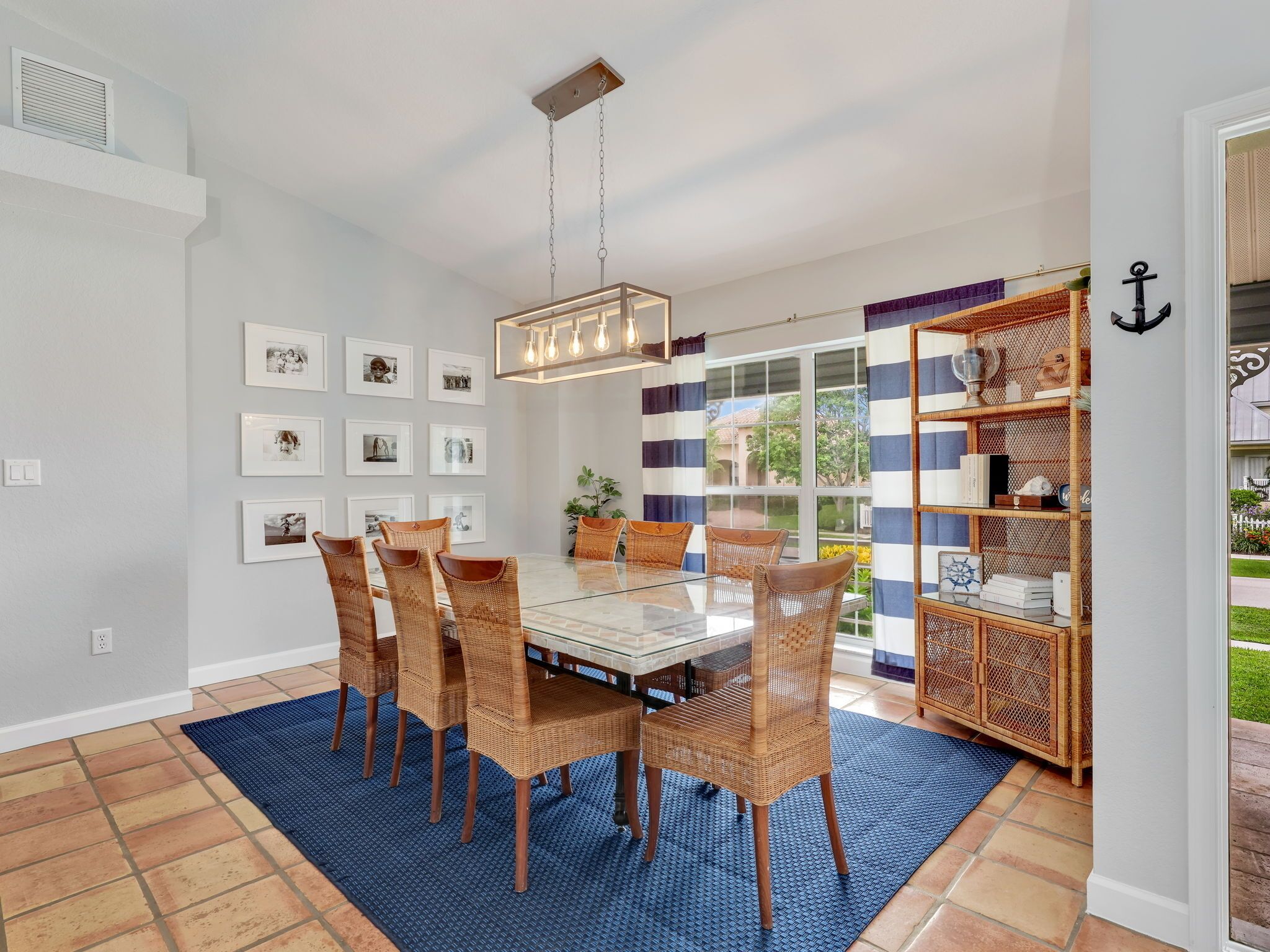 A dining area with a glass table, wicker chairs, and a blue rug. There are framed pictures on the wall and a bookshelf with various items.
