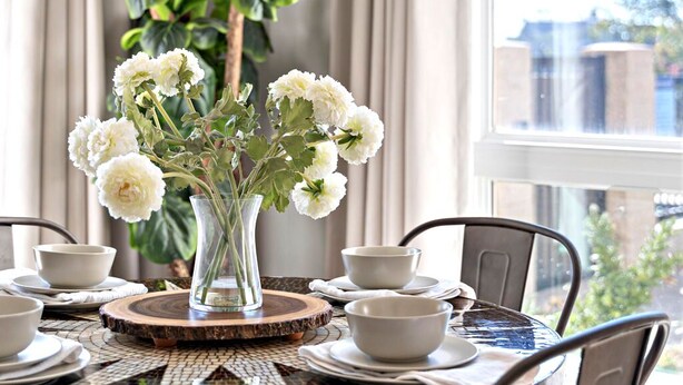 A dining area with a round table, chairs, a woven light fixture, a vase with white flowers, and a large window with curtains. - Bright, Cozy Riverfront Condo walkable to Downtown Nashville! (Nashville)