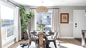A dining area with a round table, chairs, a hanging light fixture, and a large window with curtains. - Bright, Cozy Riverfront Condo walkable to Downtown Nashville! (Nashville)