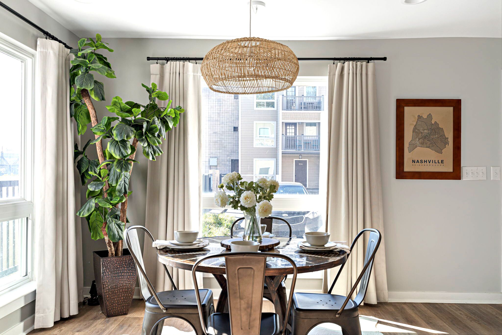 A dining area with a round wooden table, four chairs, a hanging rattan light fixture, a potted plant, and a framed map of Nashville on the wall.