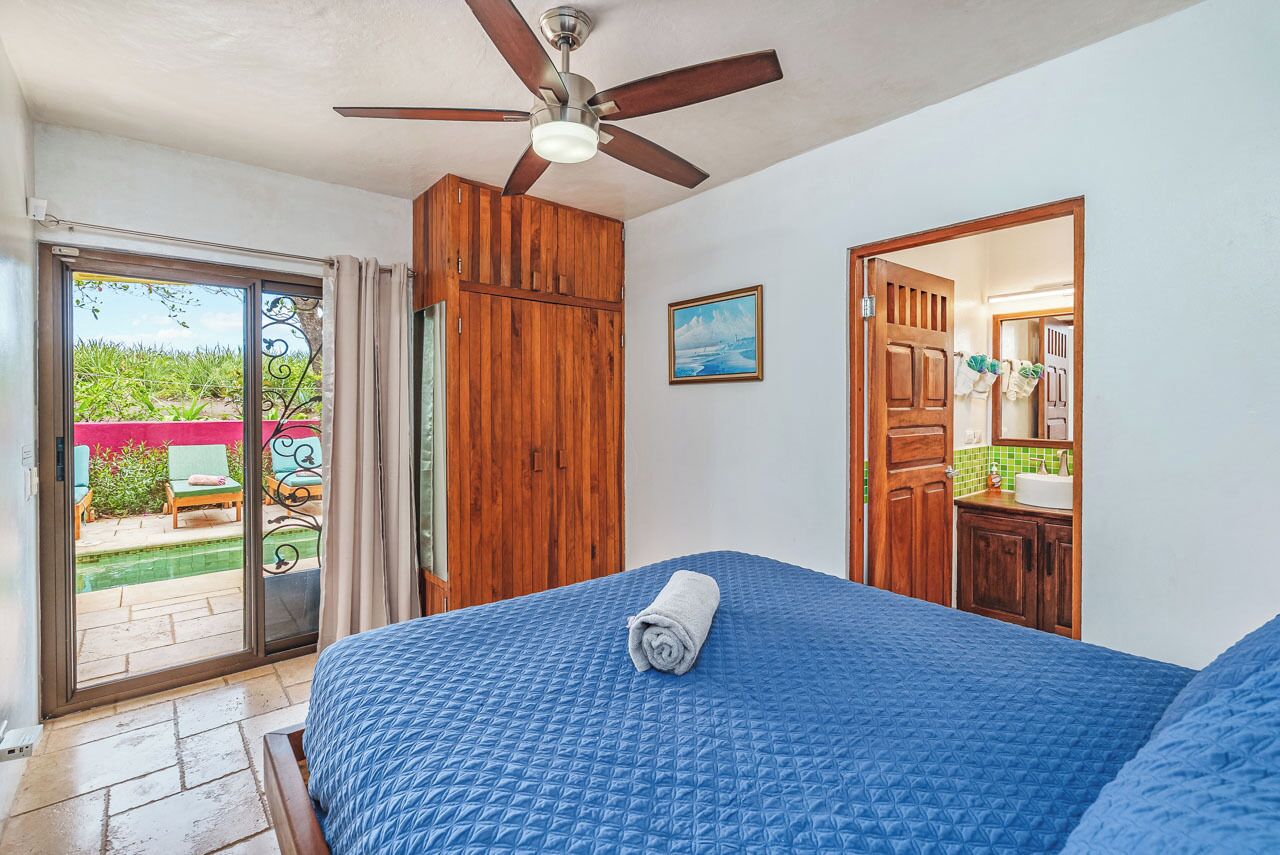 A bedroom with a blue quilted bedspread, a ceiling fan, wooden wardrobe, and a sliding glass door leading to an outdoor area.