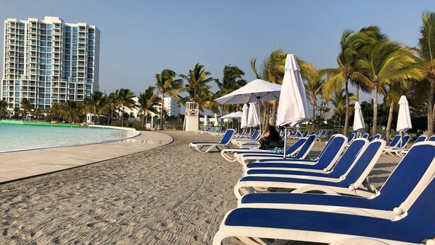 Una playa con filas de tumbonas, una piscina y palmeras.