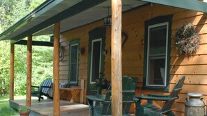 En stuga av trä med en veranda, gröna Adirondack-stolar och ett bord.
