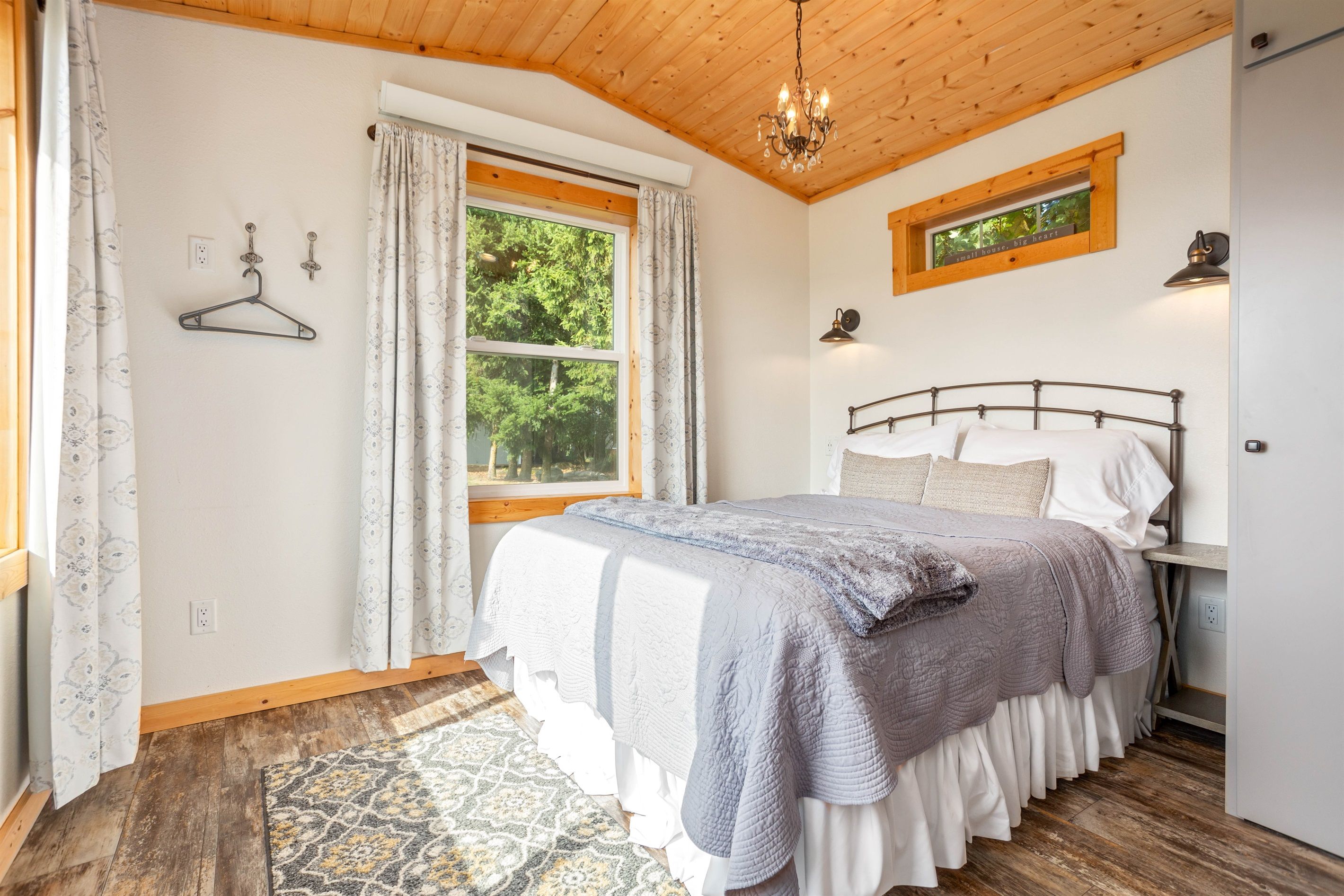 Une chambre à coucher avec un lit, un luminaire suspendu, une fenêtre avec des rideaux et une vue sur les arbres à l’extérieur.