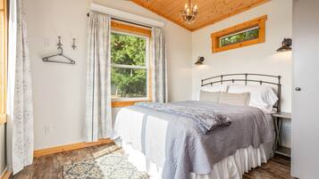 Une chambre à coucher avec un lit, un luminaire suspendu, une fenêtre avec des rideaux et une vue sur les arbres à l’extérieur.