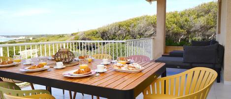 A breakfast table set with plates of food, cups, and a view of the sea.