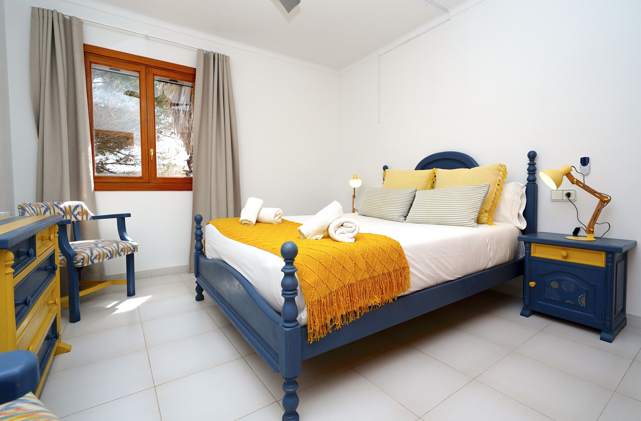 Een slaapkamer met een bed, nachtkastjes, een stoel, een dressoir en een raam met gordijnen.