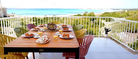 A wooden table with plates of food and drinks, surrounded by colorful chairs, on a balcony with a view of the beach and ocean.