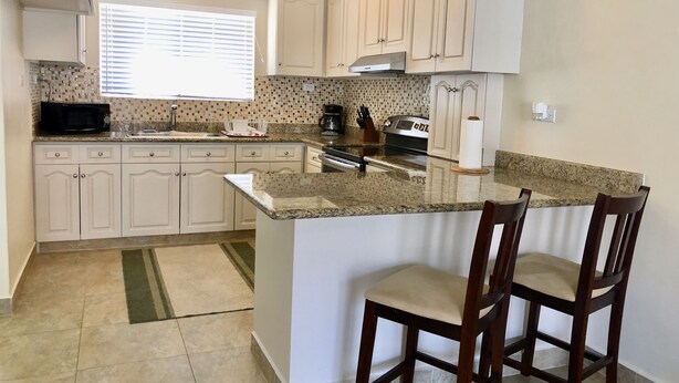 Cocina con armarios blancos, encimera de granito, microondas, estufa y dos taburetes de bar.