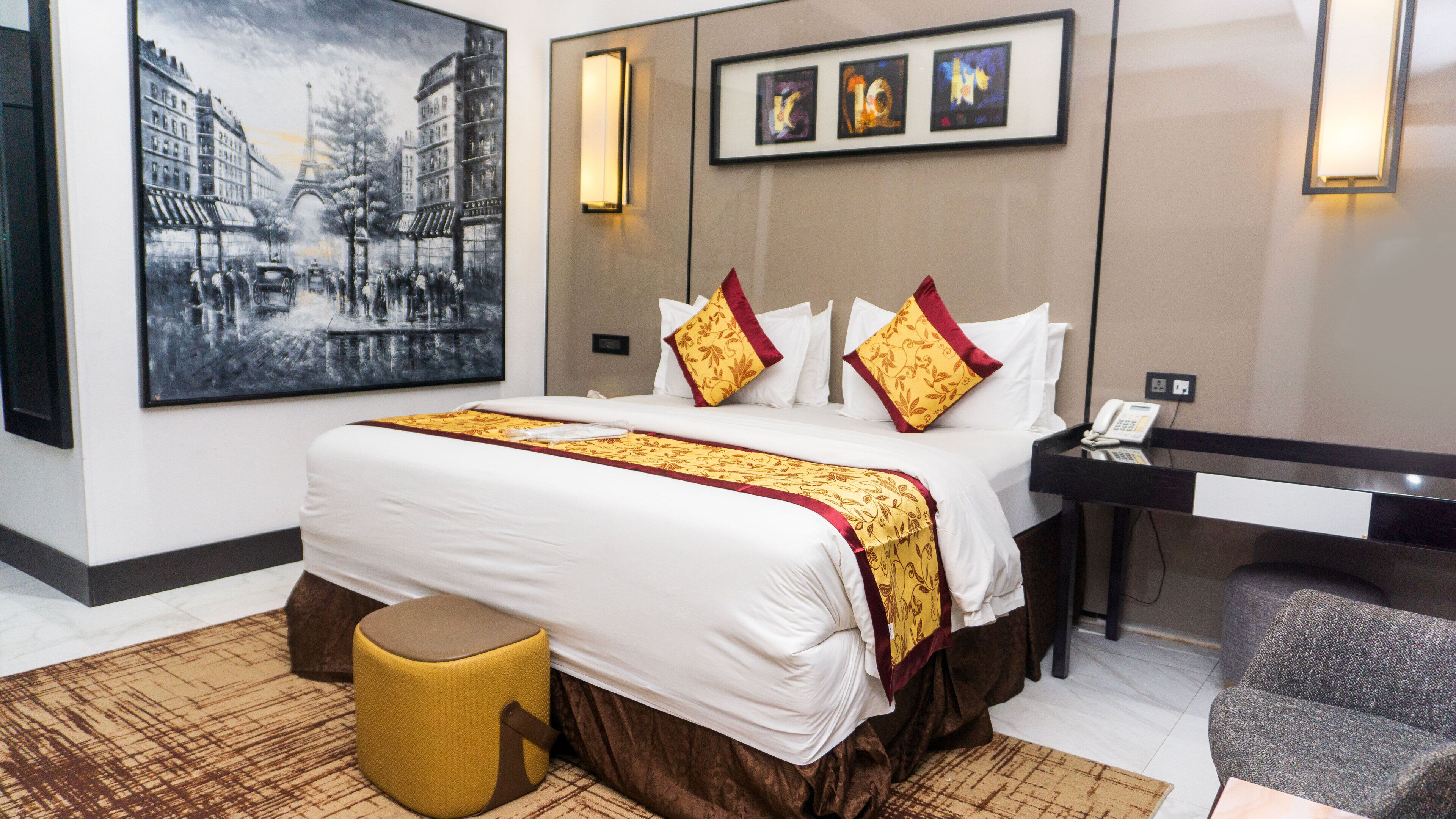 A hotel room with a large bed, a desk, a chair, a small stool, and artwork on the walls.