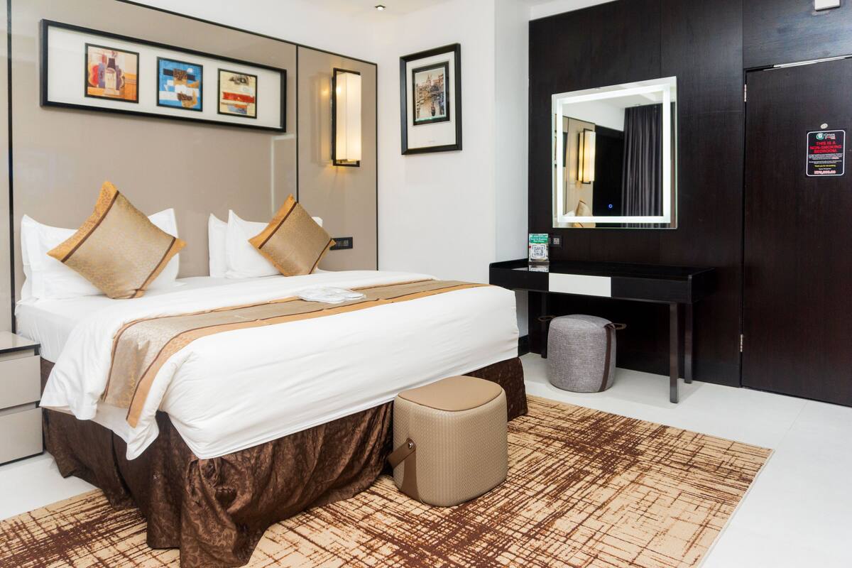 Habitación de hotel con una cama grande, mesita de noche y un espejo. Hay cuadros enmarcados en la pared y un cubo de basura junto a la cama.
