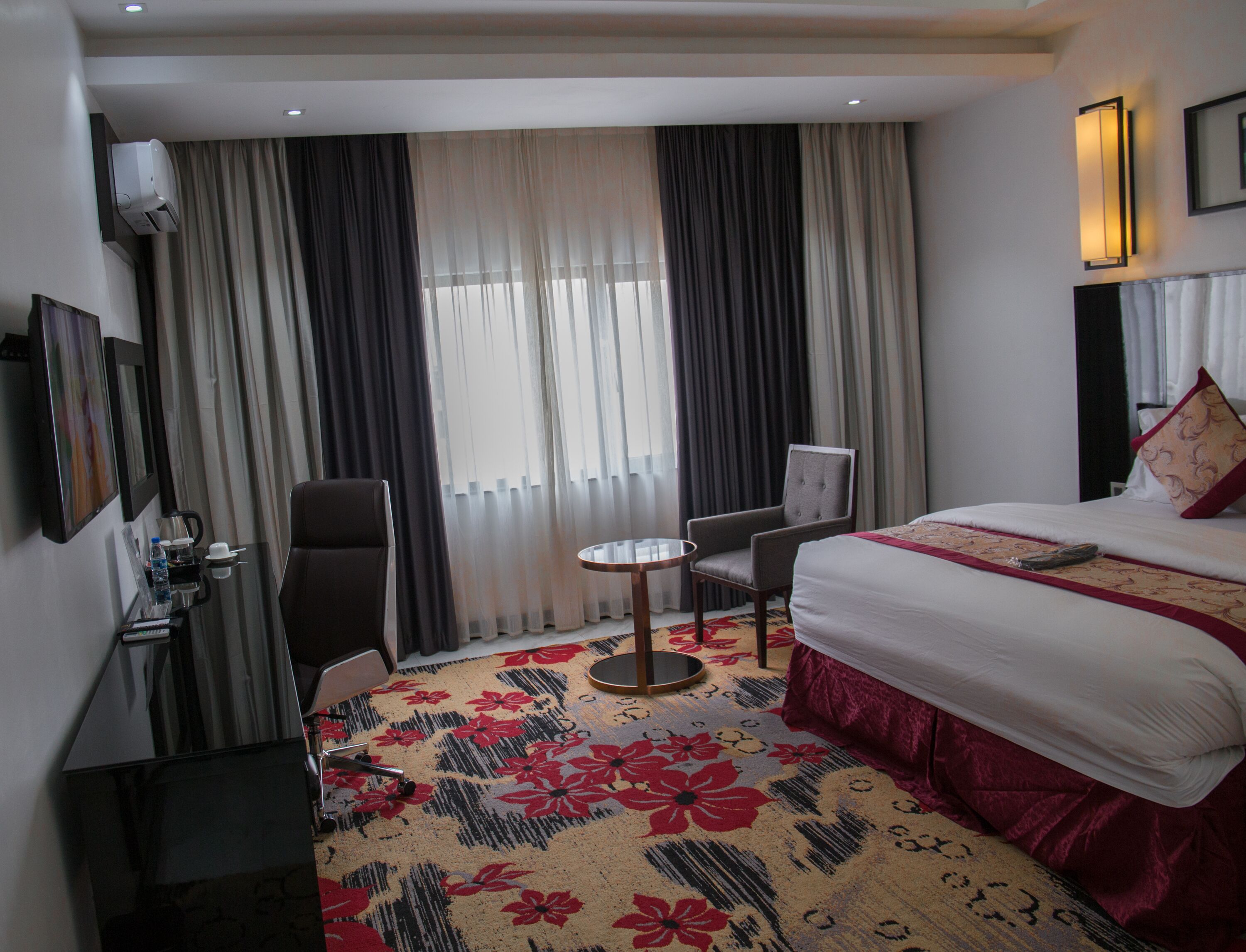 Habitación de hotel con cama, escritorio, silla, mesita pequeña y un ventanal grande con cortinas.
