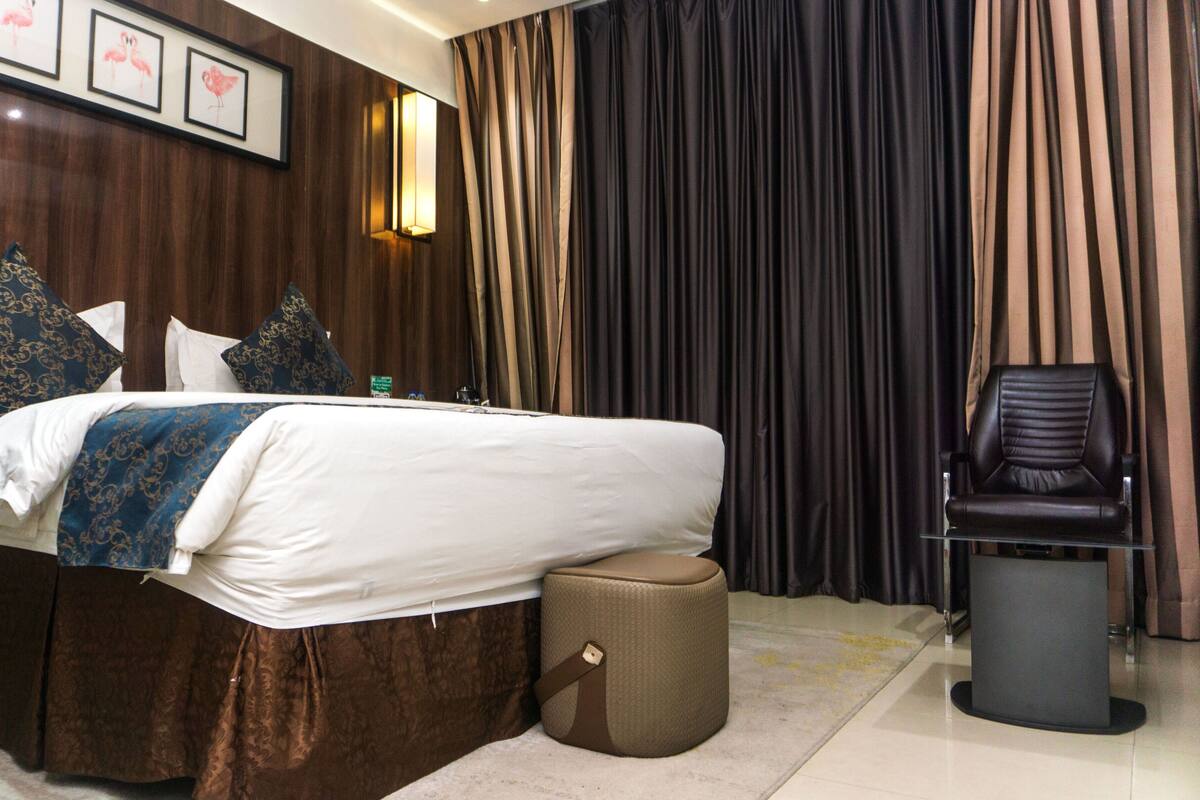 Una habitación de hotel con una cama grande, una silla, un pequeño otoman y un escritorio.
