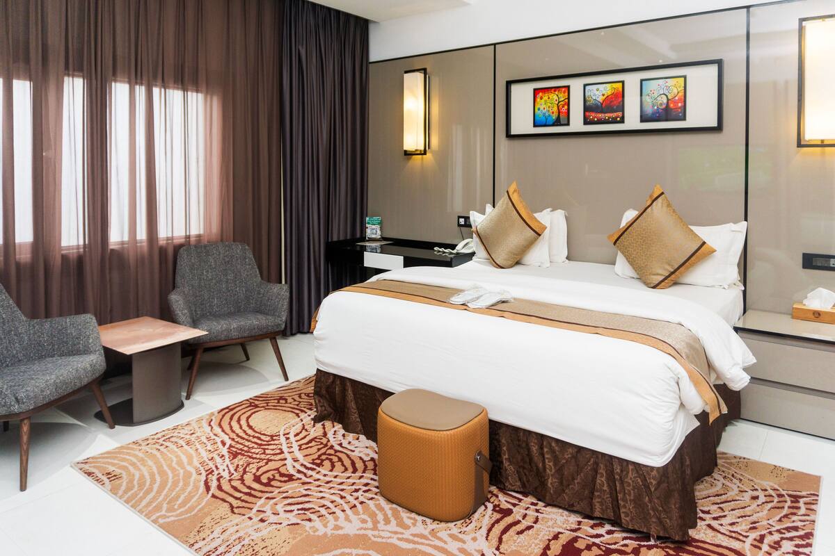 Habitación de hotel con cama, dos sillones, una mesita, una mesita de noche y un televisor instalado en la pared.