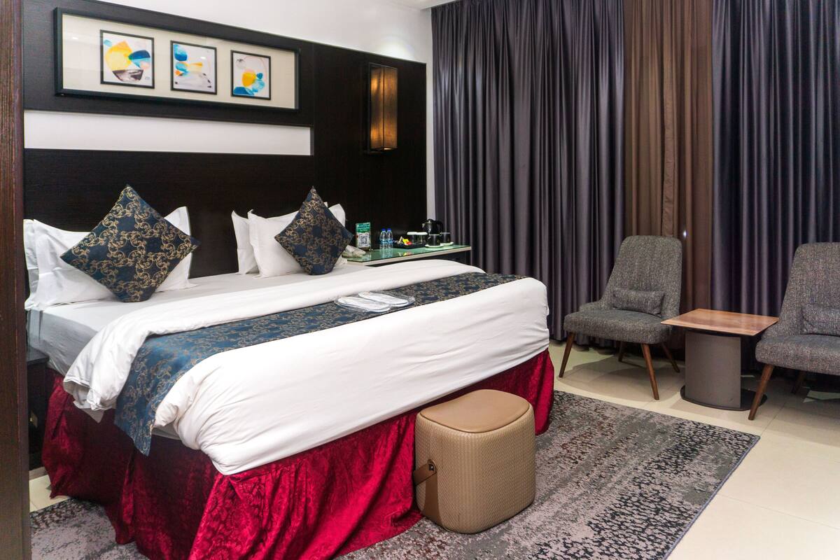 Una habitación de hotel con una cama grande, una mesita pequeña, dos sillas, una falda para la cama roja y un ventanal con cortinas.