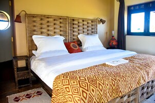 Una cama con una colcha estampada, dos almohadas blancas y una almohada roja. Hay una mesita de noche de madera con un cajón. La habitación tiene un cabecero tejido y una ventana con cortinas.