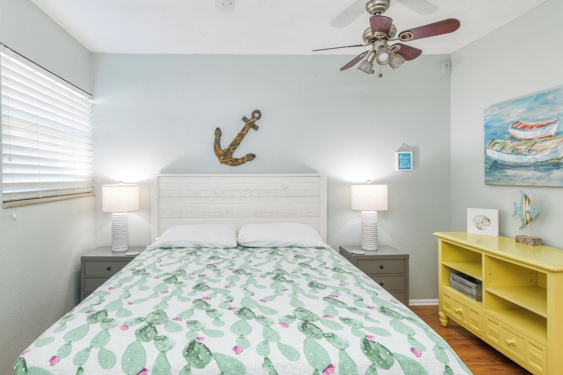 A bedroom with a bed, two bedside lamps, a ceiling fan, a wall anchor decoration, a small framed picture, and a yellow dresser.