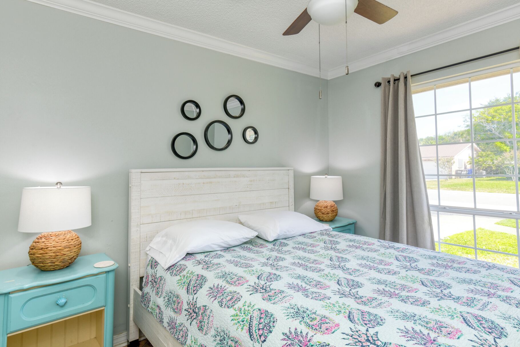A bedroom with a bed, two bedside tables, a ceiling fan, a window with curtains, and wall-mounted mirrors.
