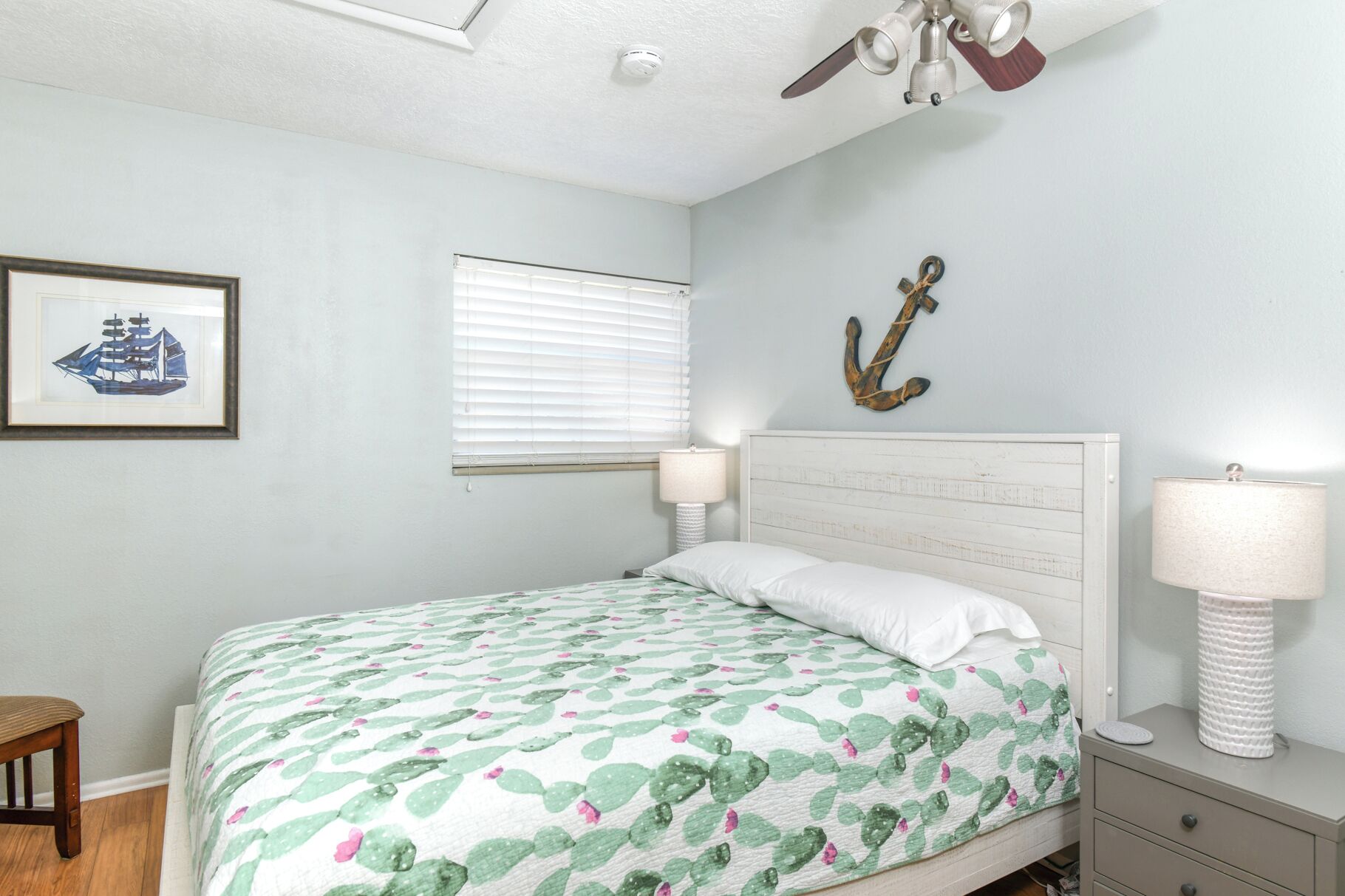 A bedroom with a bed, two bedside lamps, a ceiling fan, a framed picture, a window with blinds, and a wall anchor decoration.