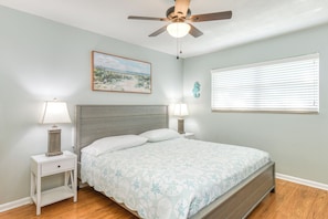 A bedroom with a bed, two bedside tables with lamps, a ceiling fan, a window with blinds, and a wall-mounted seahorse decoration.