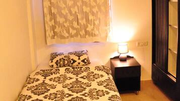 A bedroom with a bed, a nightstand, a lamp, a window with curtains, and a wardrobe.