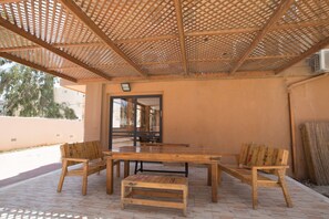An outdoor patio with a wooden table and benches, a pergola overhead, and a view of the street and buildings in the background.