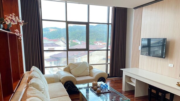 A living room with a sofa, a glass coffee table, a TV, and a view of buildings through large windows. - Anoulackkhenlao Hotel (Phonsavan)