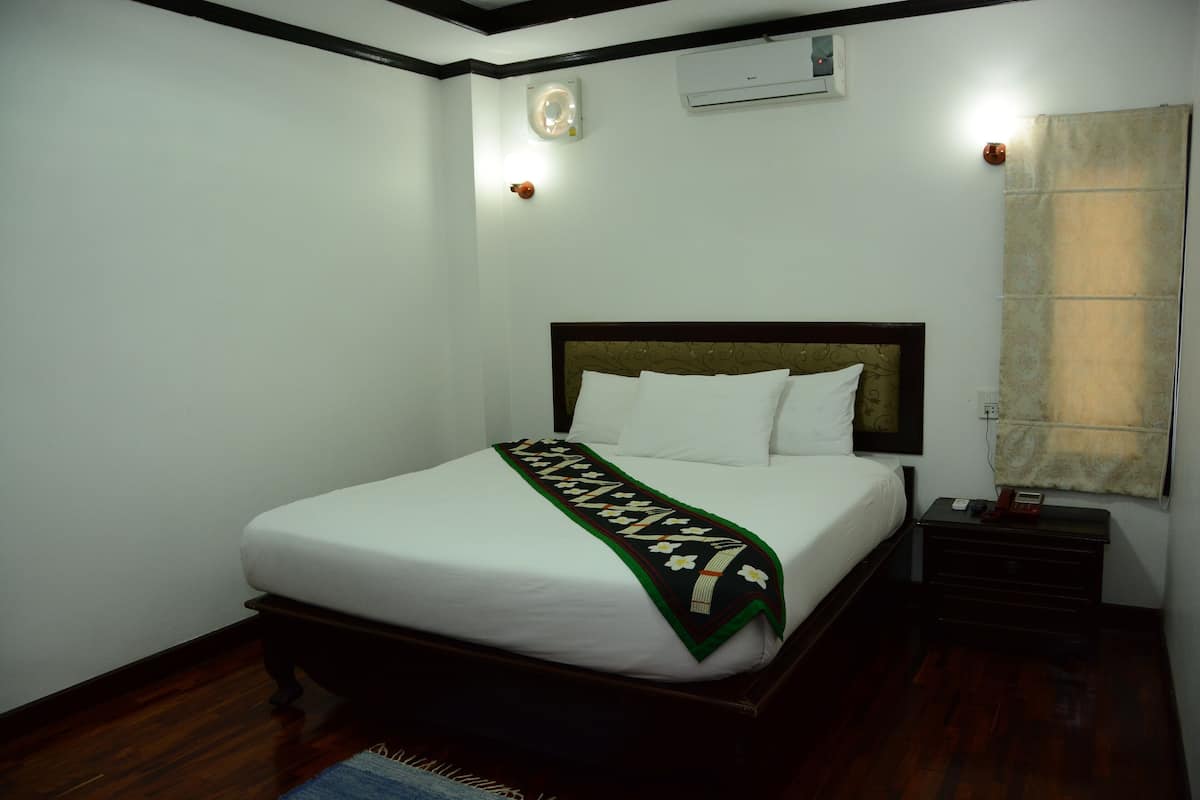 A neatly made bed with a white comforter and a green decorative throw. There is a wooden nightstand with a lamp and a telephone. The room has white walls and a wooden floor.