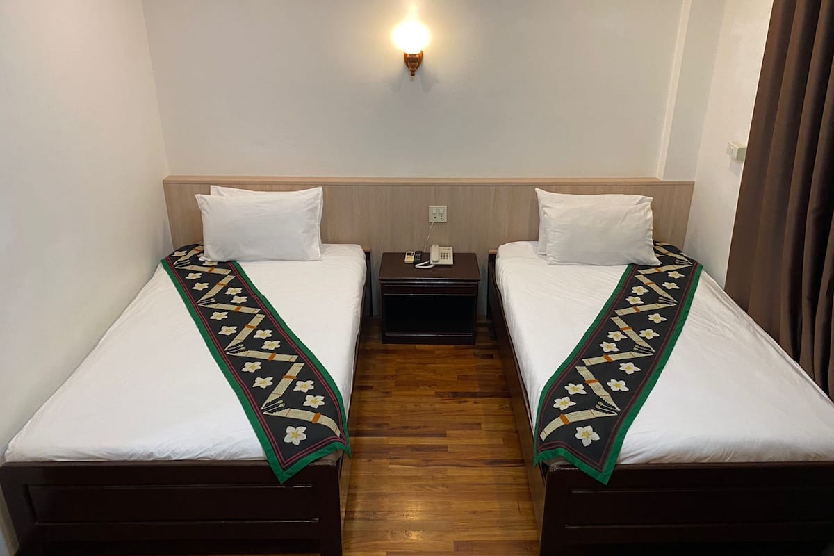 A hotel room with two single beds, each with a white pillow and a patterned bedspread. There is a nightstand between the beds with a phone and a lamp. The room has a wooden floor and a wall-mounted light fixture above the beds.