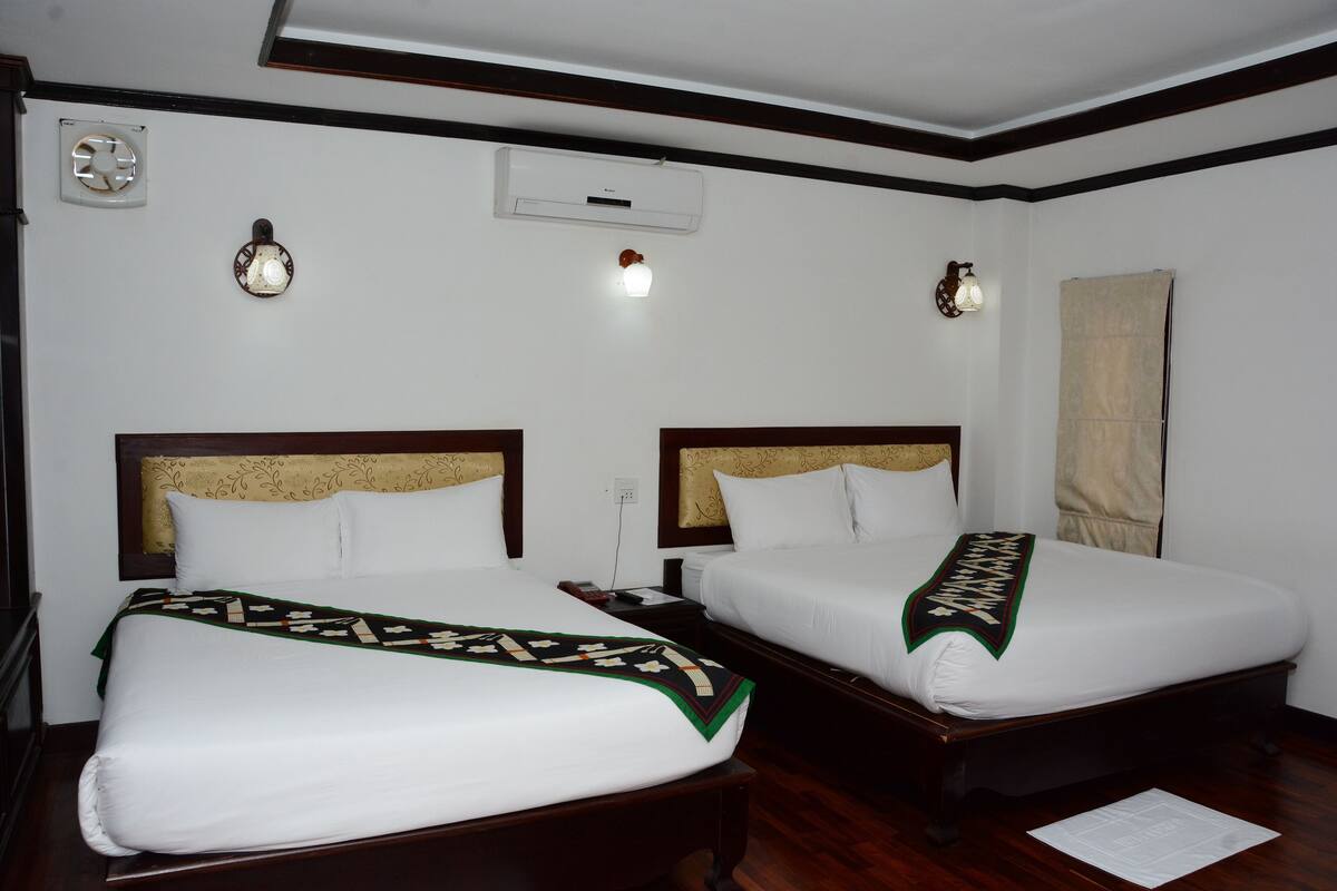 A hotel room with two beds, a ceiling fan, a wall-mounted air conditioner, and a nightstand with a lamp.