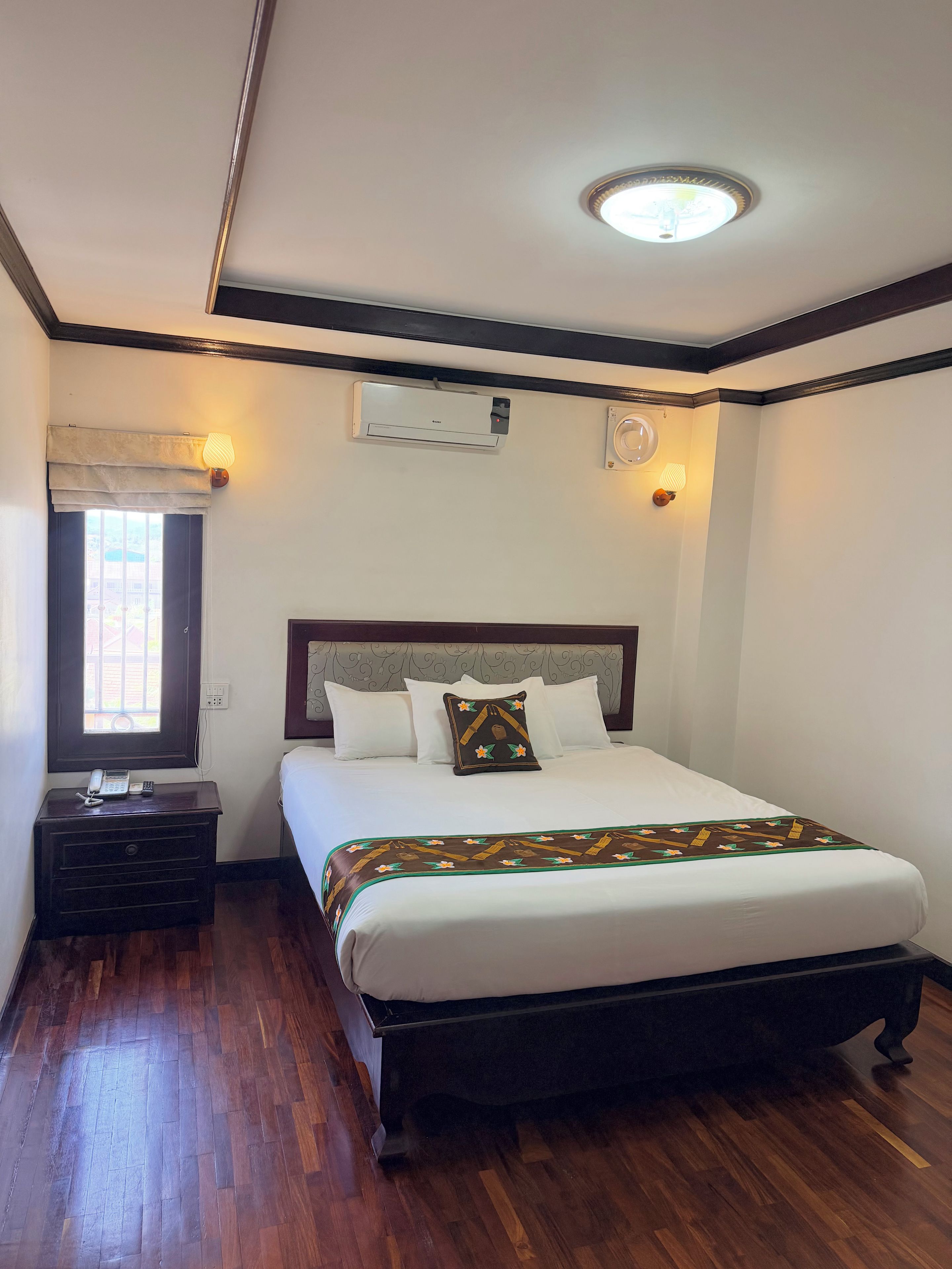 A hotel room with a bed, bedside tables, a window with curtains, an air conditioning unit, and a wall-mounted light fixture.