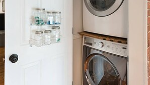 A laundry room with a front-loading washing machine and dryer, a white door, and a shelf with jars. - The Grey Goose (Waterfront) Historic New Bern - Private Beach - BRING YOUR BOAT! (New Bern)