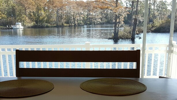 A table with two woven placemats and a wooden chair with a backrest, set against a view of a lake surrounded by trees. - The Grey Goose (Waterfront) Historic New Bern - Private Beach - BRING YOUR BOAT! (New Bern)