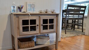 A wooden console table with glass doors, a wicker basket, and a stack of books. - The Grey Goose (Waterfront) Historic New Bern - Private Beach - BRING YOUR BOAT! (New Bern)
