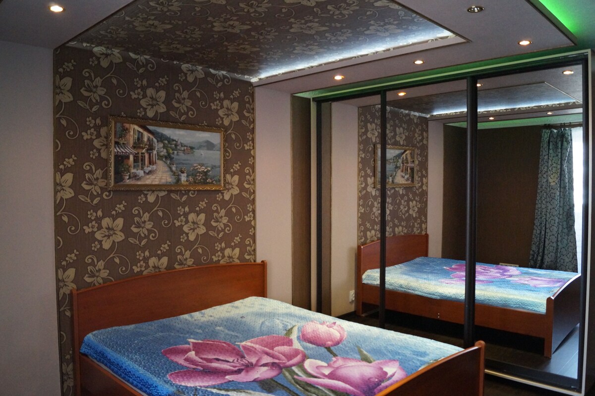 A room with two wooden beds, floral wallpaper, and a large mirror reflecting the room.