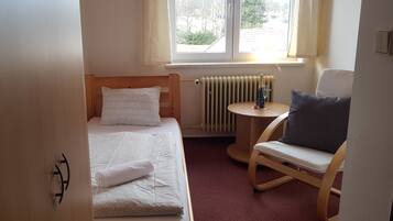 A small, neatly arranged room with a single bed, a chair, a small table, and a window providing natural light.