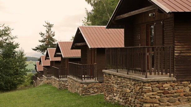 A row of log cabins with stone foundations and metal roofs, set in a grassy area with trees in the background. - Hotel Rohanov (Vacov)