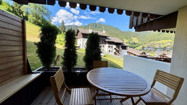 A wooden deck with a round table and two chairs, overlooking a village with houses and a mountain. - Cozy studio 4 pers, ski-in/ski-out, terrace, garage, WIFI (La Clusaz)