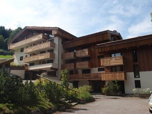 A multi-story building with wooden balconies, a ski slope visible in the background, and a parking area in front. - Cozy studio 4 pers, ski-in/ski-out, terrace, garage, WIFI (La Clusaz)