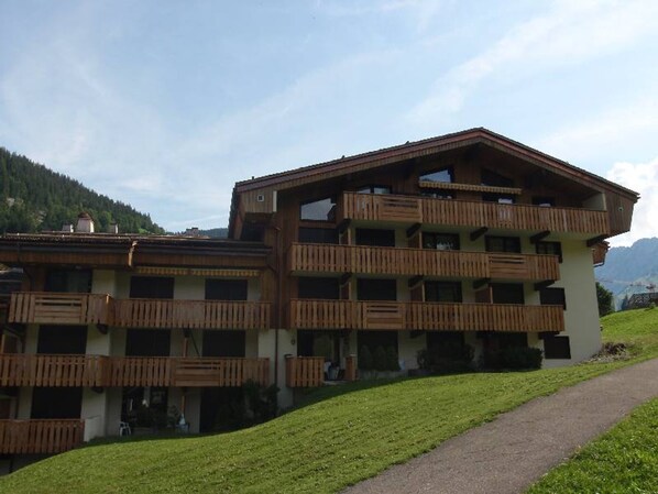 A multi-story building with wooden balconies, a paved path, and a mountainous backdrop. - Cozy studio 4 pers, ski-in/ski-out, terrace, garage, WIFI (La Clusaz)