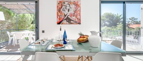 A modern dining area with a glass table, white chairs, and a painting above.