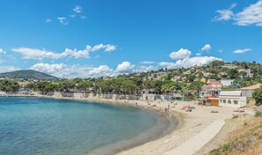 A coastal town with a sandy beach, clear blue sea, and buildings on a hillside.