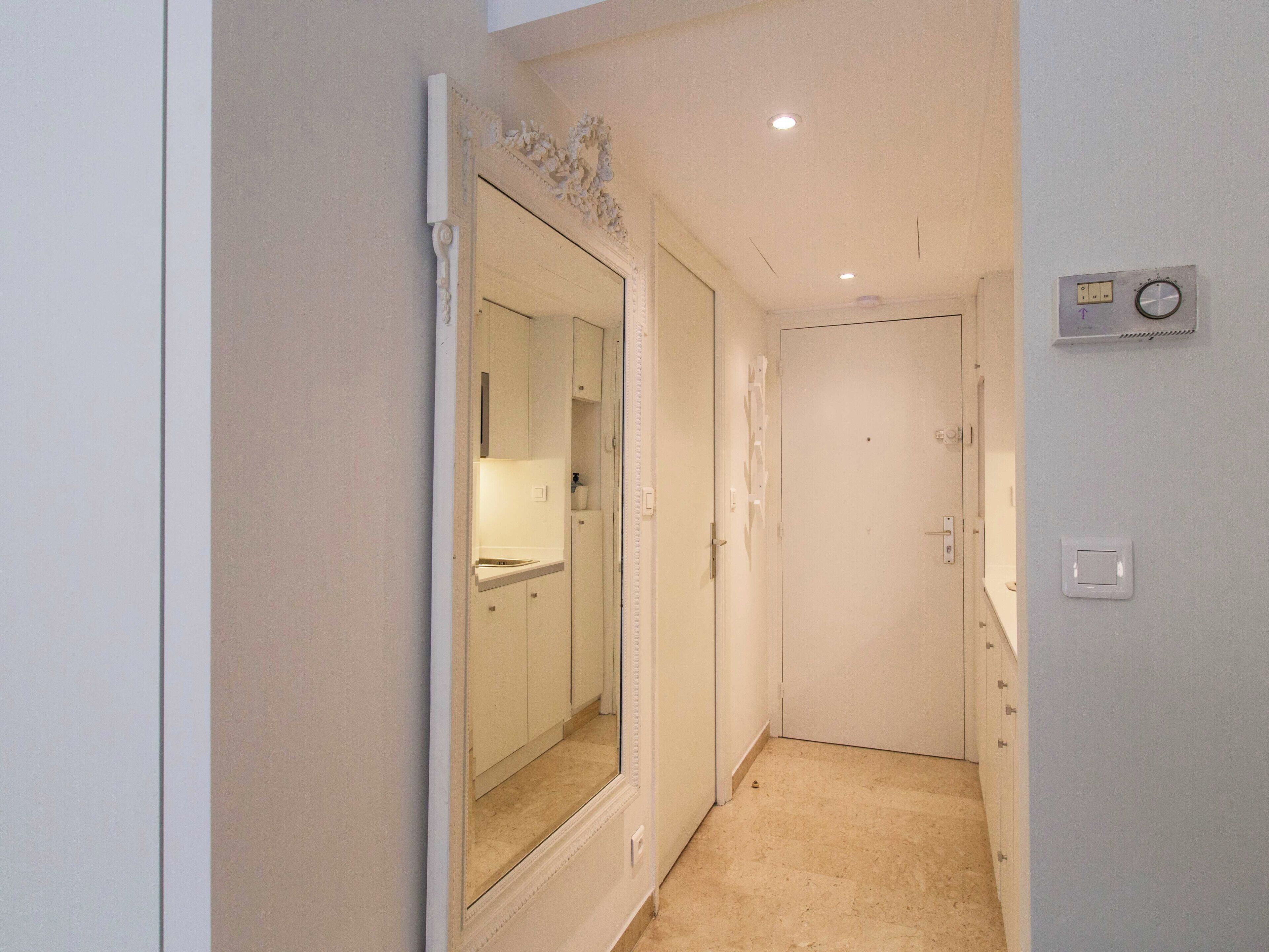 A narrow hallway with a door at the end, a mirror on the left, and a thermostat on the wall.