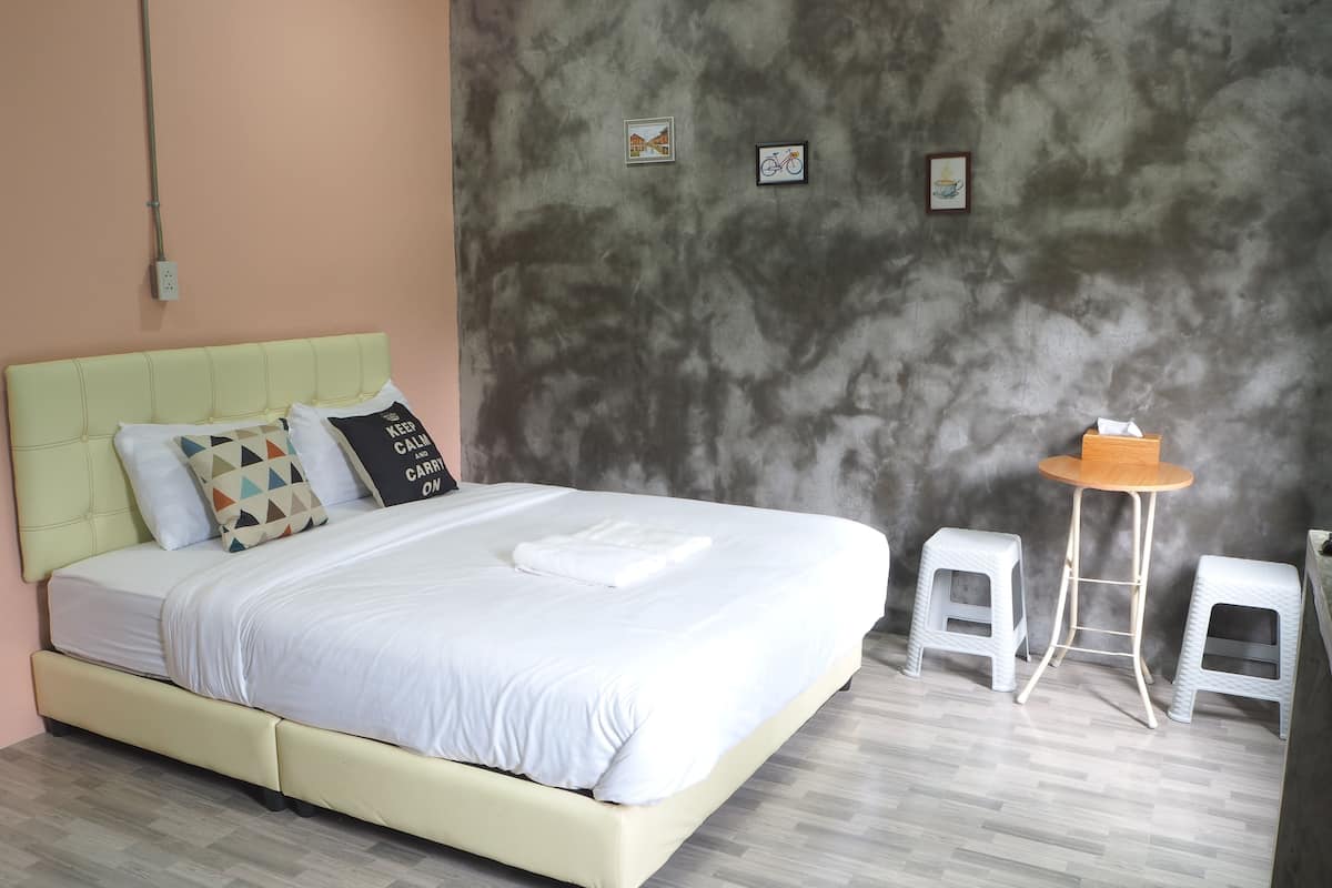 Una cama con cabecera verde claro, ropa de cama blanca y varias almohadas. Junto a la cama hay una pequeña mesa de madera con un taburete. La habitación tiene una pared de concreto con dos cuadros enmarcados y un enchufe.